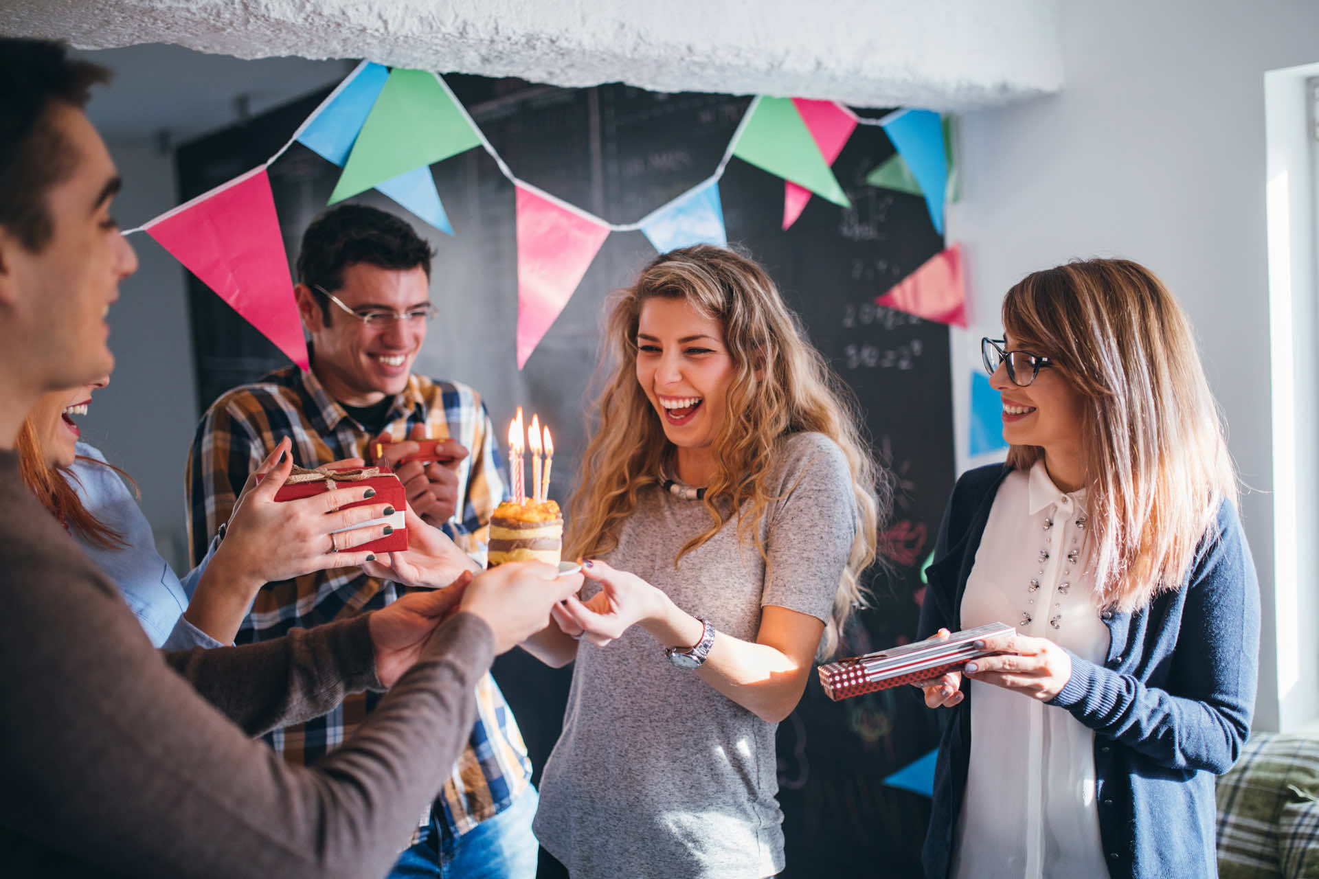 A group of friends celebrating with a birthday cake and gifts in a decorated room with colorful bunting.