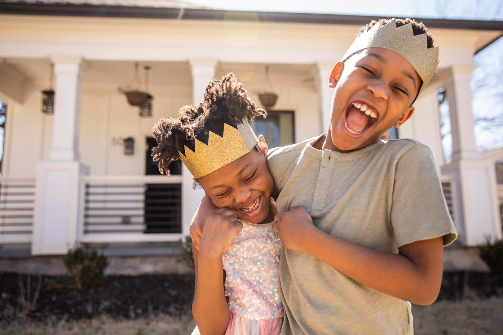 Two children are smiling and playing outside in front of a house. One child is wearing a golden crown and a shiny dress