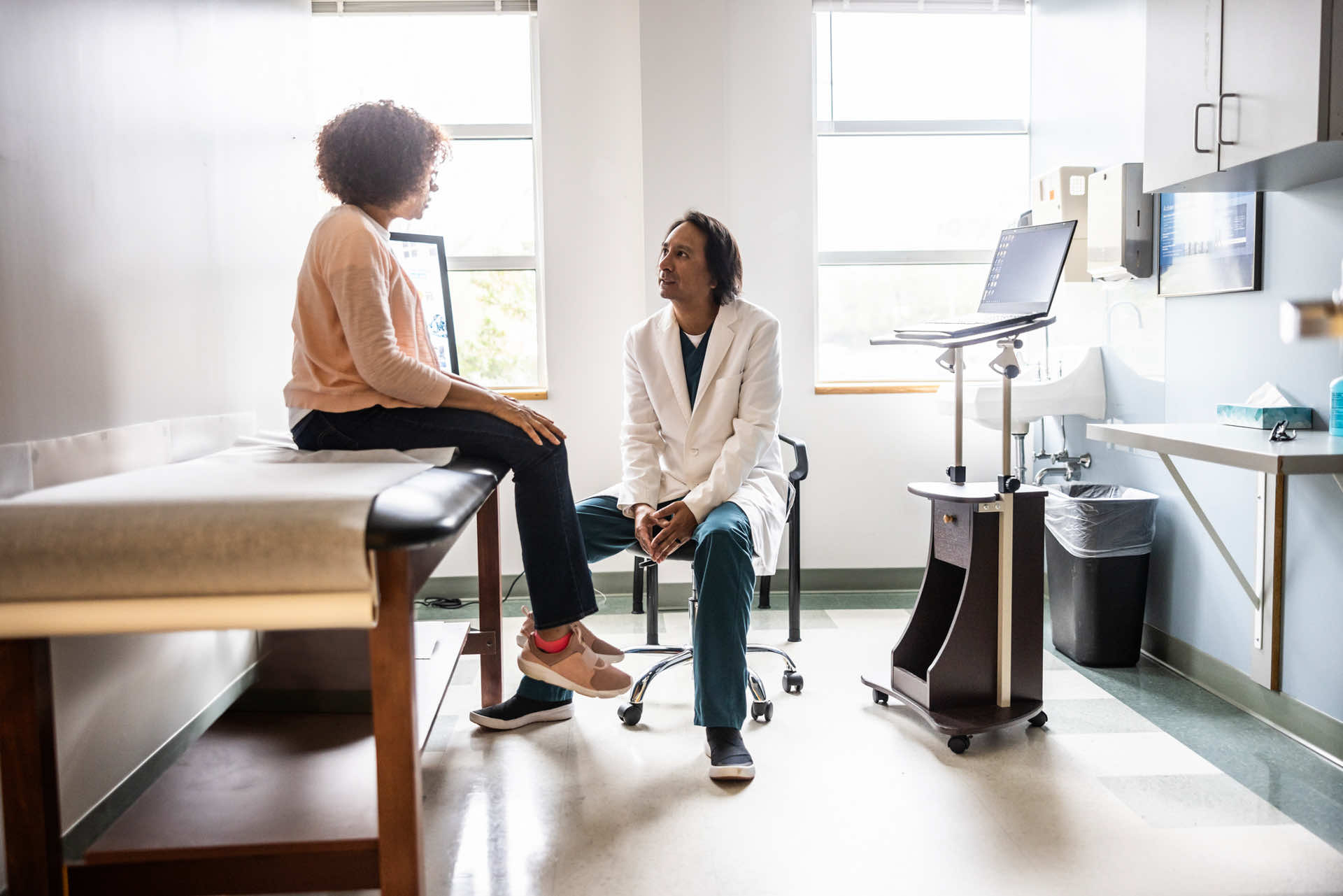 A doctor in a white coat talking to a patient sitting on an exam table in a medical office with natural light coming through the windows