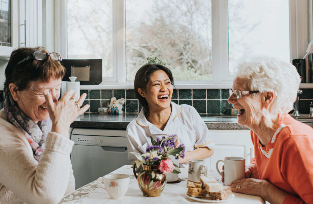 Three women sitting at a table laughing together with cups of tea and a flower vase in front of them